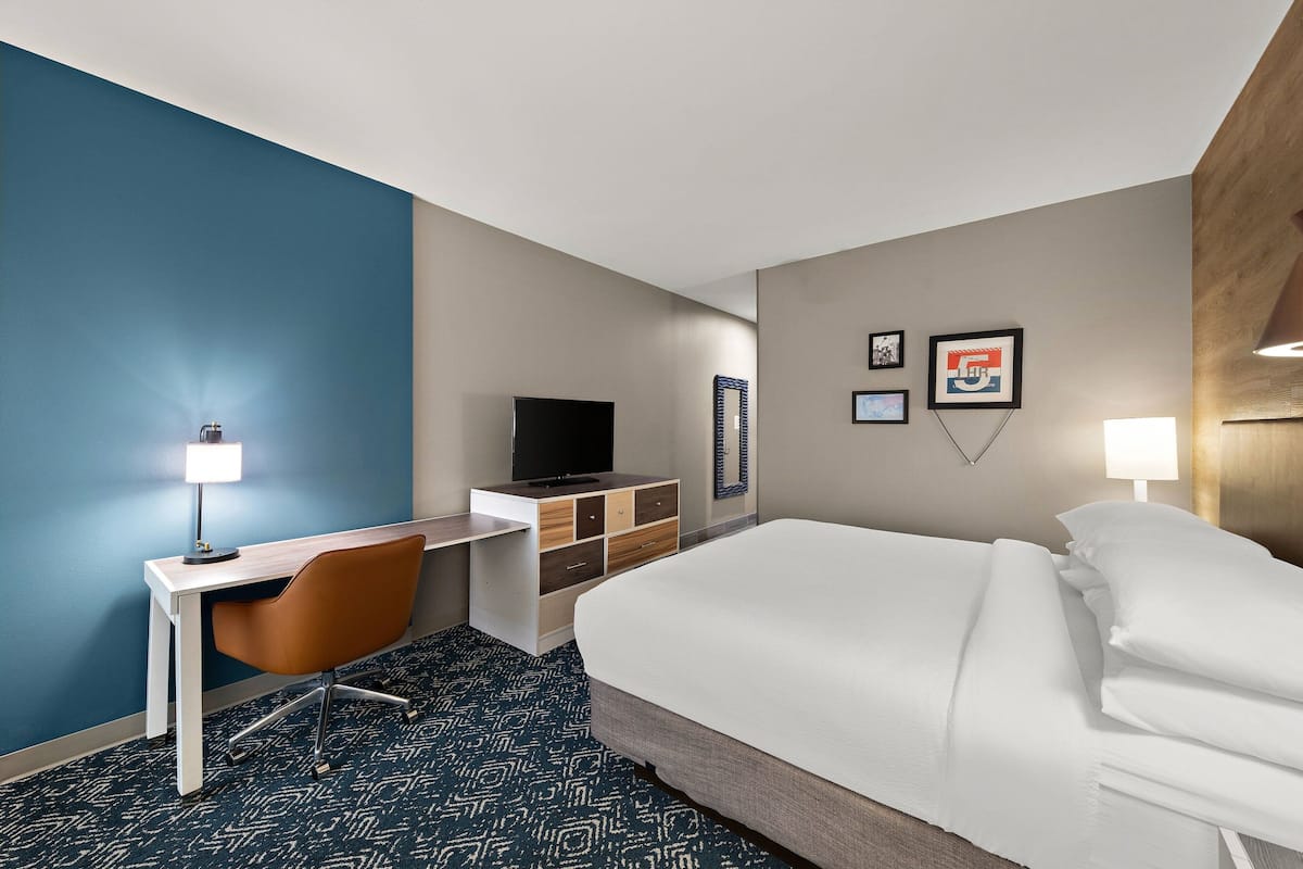 A hotel room with a bed, desk, chair, TV, and wall art.