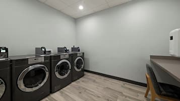 A laundry room with two front-loading washers and dryers, a wooden chair, and a small table with a soap dispenser.