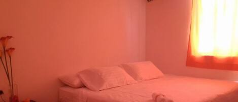A single bed with white bedding and pillows, a nightstand with a vase of flowers, and a rolled towel on the bed.