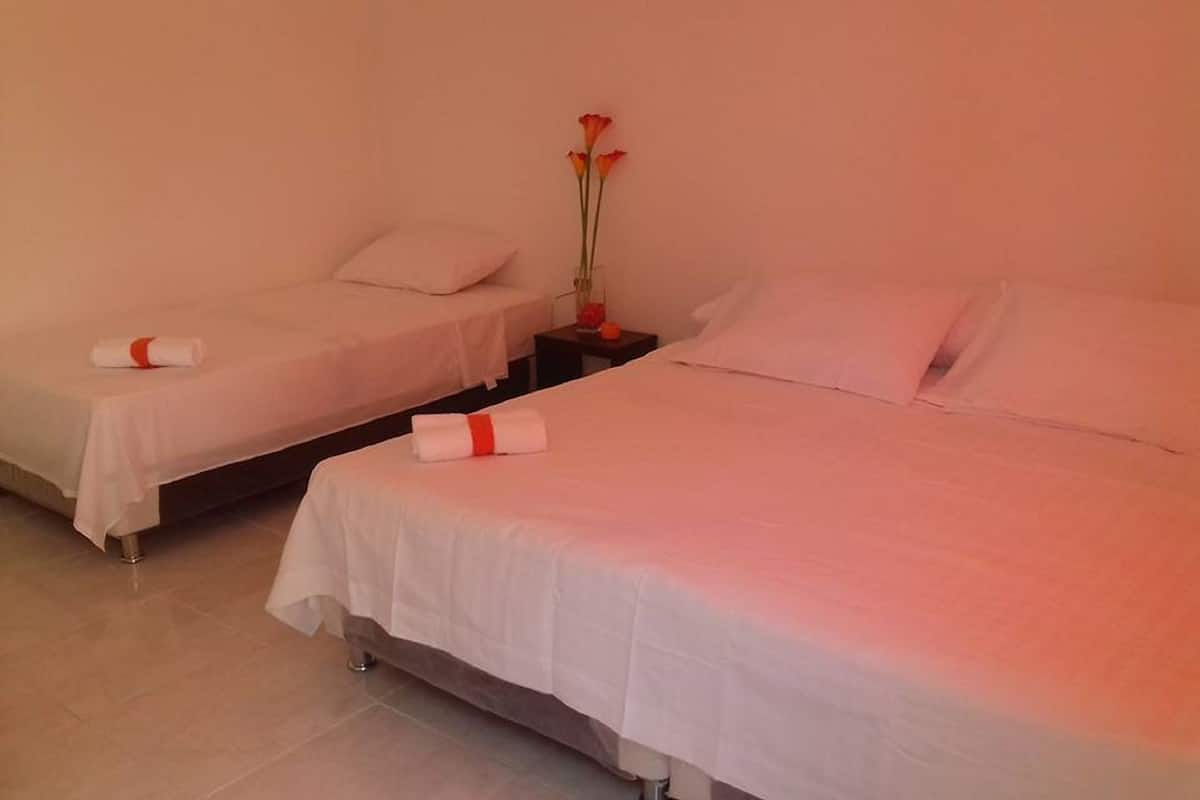 Quarto de hotel com duas camas, cada uma com roupa de cama branca e um laço vermelho. Há um vaso com flores em uma mesa de cabeceira entre as camas.