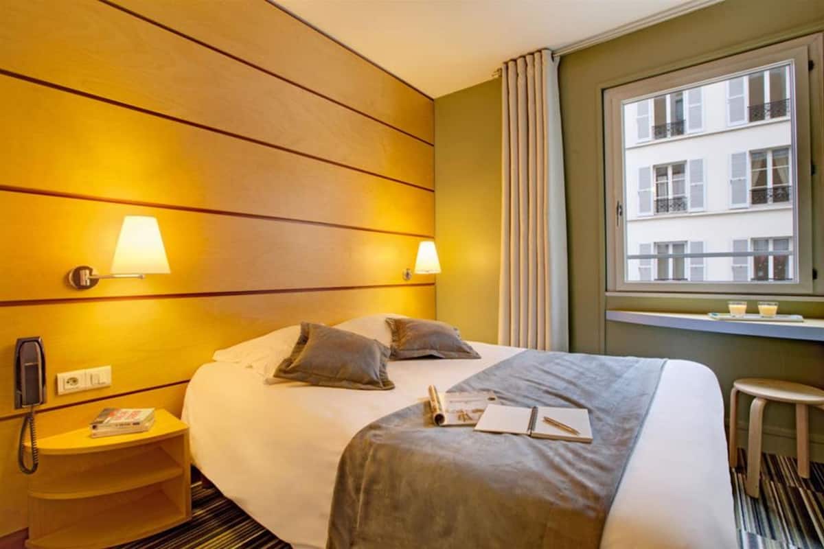 A hotel room with a bed, bedside lamps, a telephone, a small table with a book, and a window with a view of buildings.