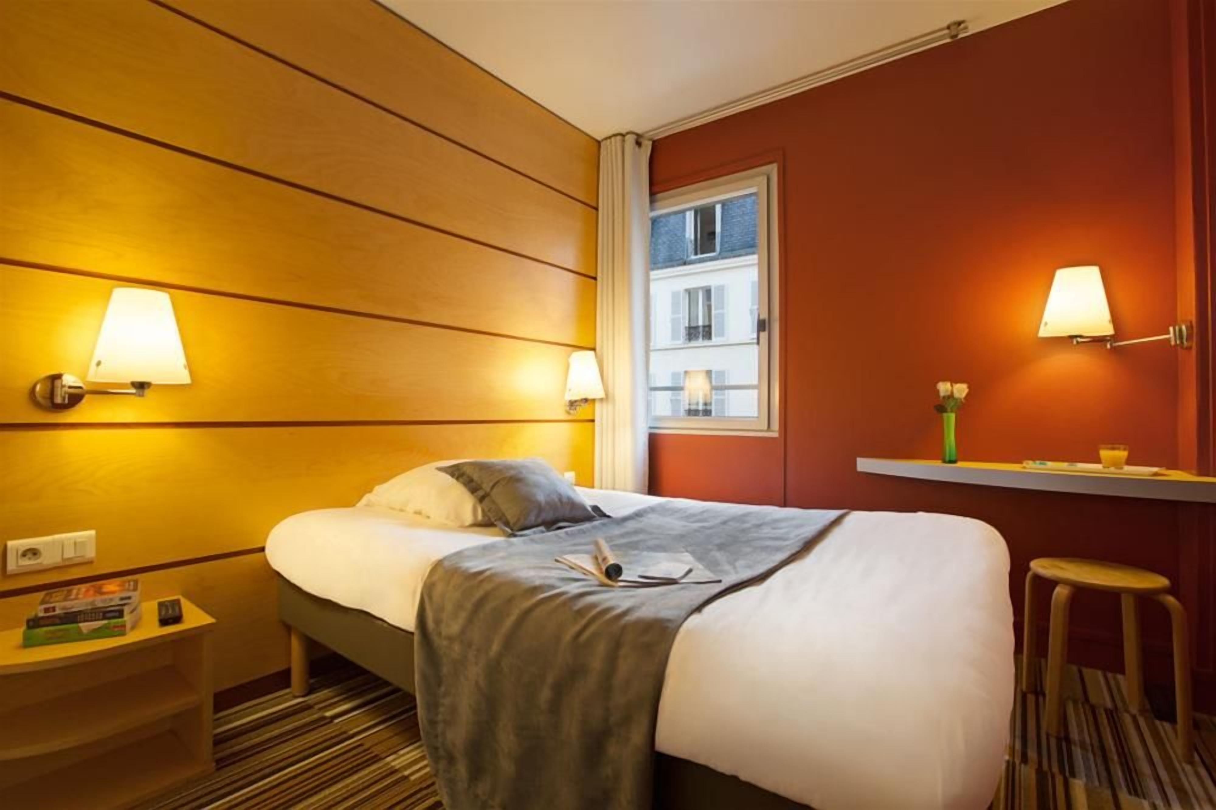 Ein Hotelzimmer mit Bett, Nachttischlampen, einem kleinen Tisch mit einem Buch, einem Fenster mit Blick auf Gebäude und einem Hocker.