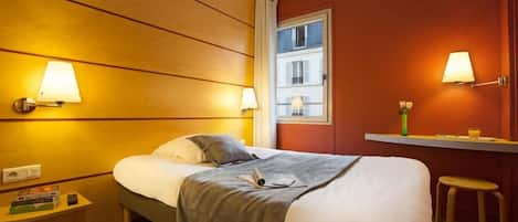 Habitación de hotel con cama, lámparas de noche, una mesita con un libro, una ventana con vistas a edificios y un taburete.