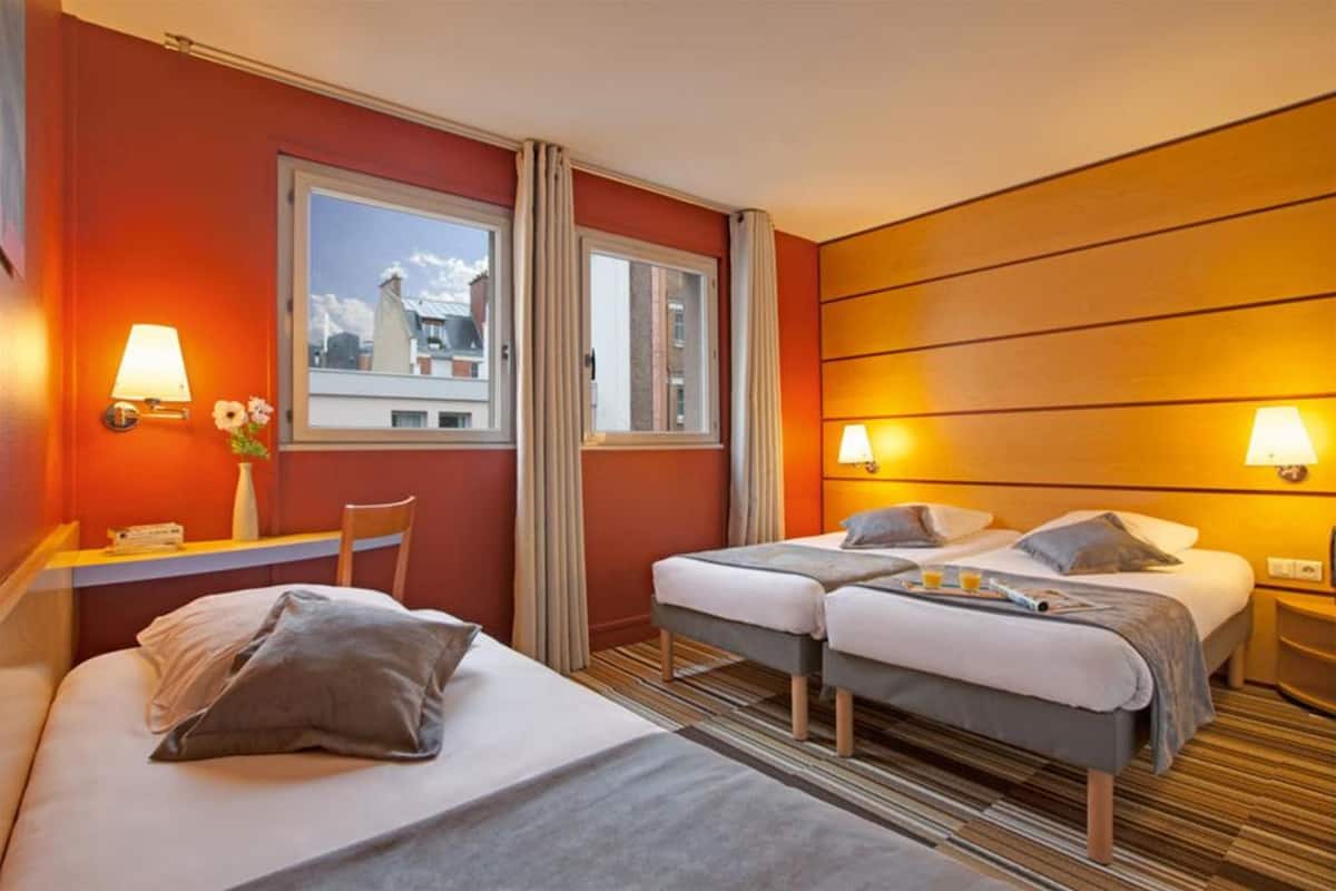 A hotel room with two beds, a desk, and a window with a view of buildings.