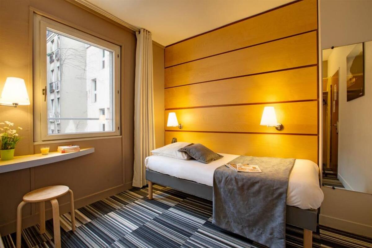 A hotel room with a bed, bedside lamps, a window with curtains, a small table with a plant, and a striped carpet.