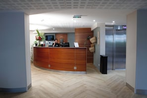 A reception desk with a receptionist, a flat-screen TV, and a digital clock. - Le Magendie (Paris)