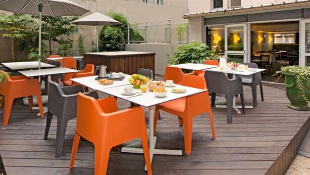 Un área de comedor al aire libre con sillas anaranjadas y mesas blancas, rodeada de follaje y una terraza de madera.