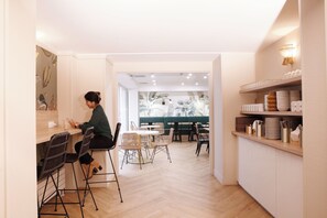 A modern interior with a dining area featuring a mix of metal and wicker chairs, a counter with a person working, and a view into a room with a green couch and a tropical mural. - Le Magendie (Paris)