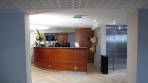 A reception desk with a receptionist, a flat-screen TV, and a digital clock. - Le Magendie (Paris)