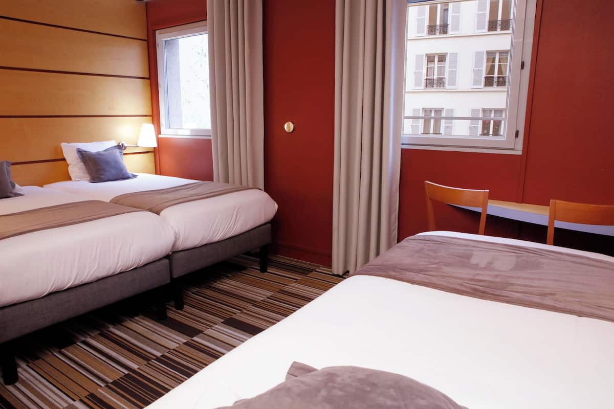 A hotel room with two beds, a wooden headboard, a window with curtains, a desk, and a chair.