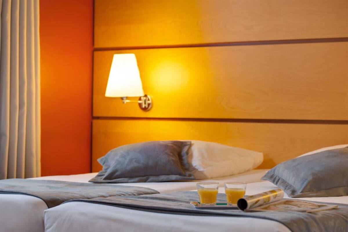 A hotel room with a wooden headboard, a bedside lamp, a bed with grey bedding, and a tray with two glasses of orange juice and a magazine.
