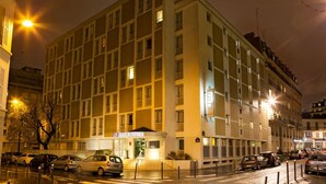 Un hotel de varios pisos con entrada central, letrero visible y área de estacionamiento con automóviles.