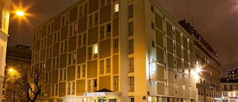 Un hotel de varios pisos con entrada central, letrero visible y área de estacionamiento con automóviles.