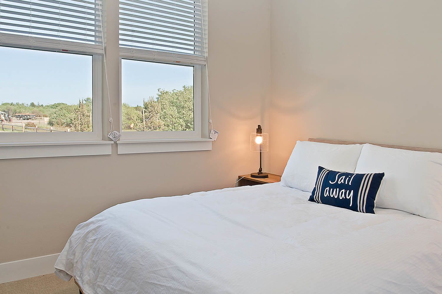 Una cama bien hecha con ropa blanca y una sola almohada con texto, una mesita de noche con lámpara, y una ventana con persianas que da a un paisaje.