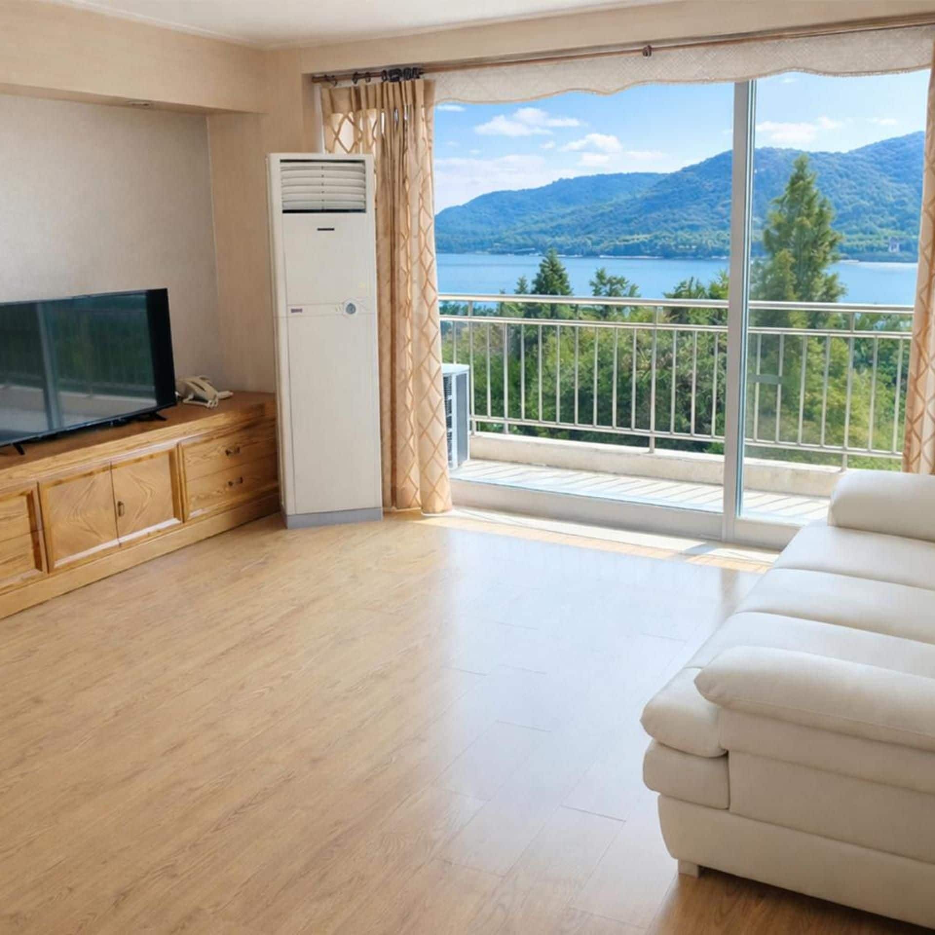 A room with a television, a white air conditioner, a wooden cabinet, a white sofa, and a balcony with a view of mountains and a lake.