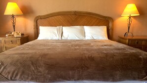 A large bed with a brown cover, white pillows, and a white ruffled skirt. There are two bedside lamps and a telephone on a wooden nightstand.