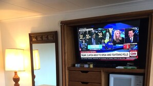 A hotel room with a wooden desk, a television displaying news, a lamp, and a microwave.