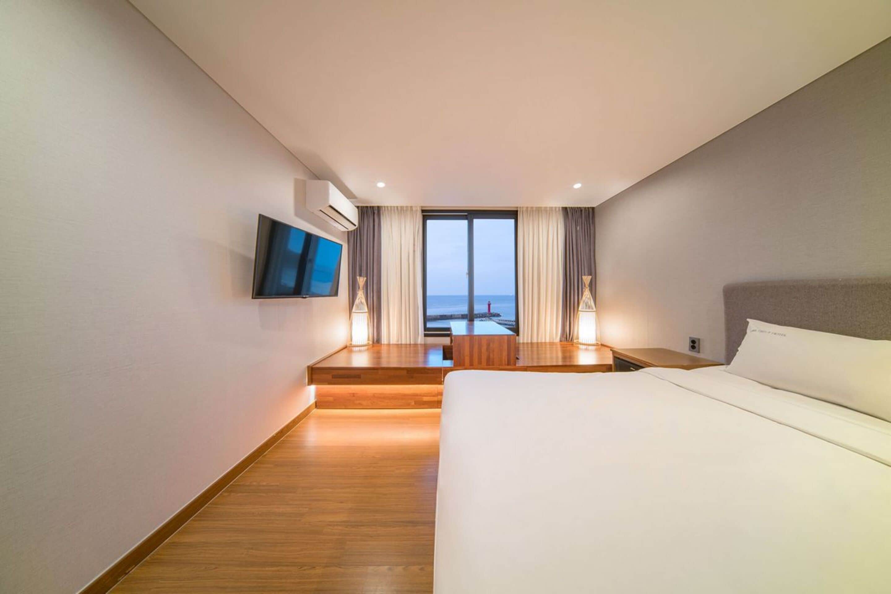 A modern hotel room with a large bed, a TV mounted on the wall, a desk with a chair, and a view of the sea through the window.
