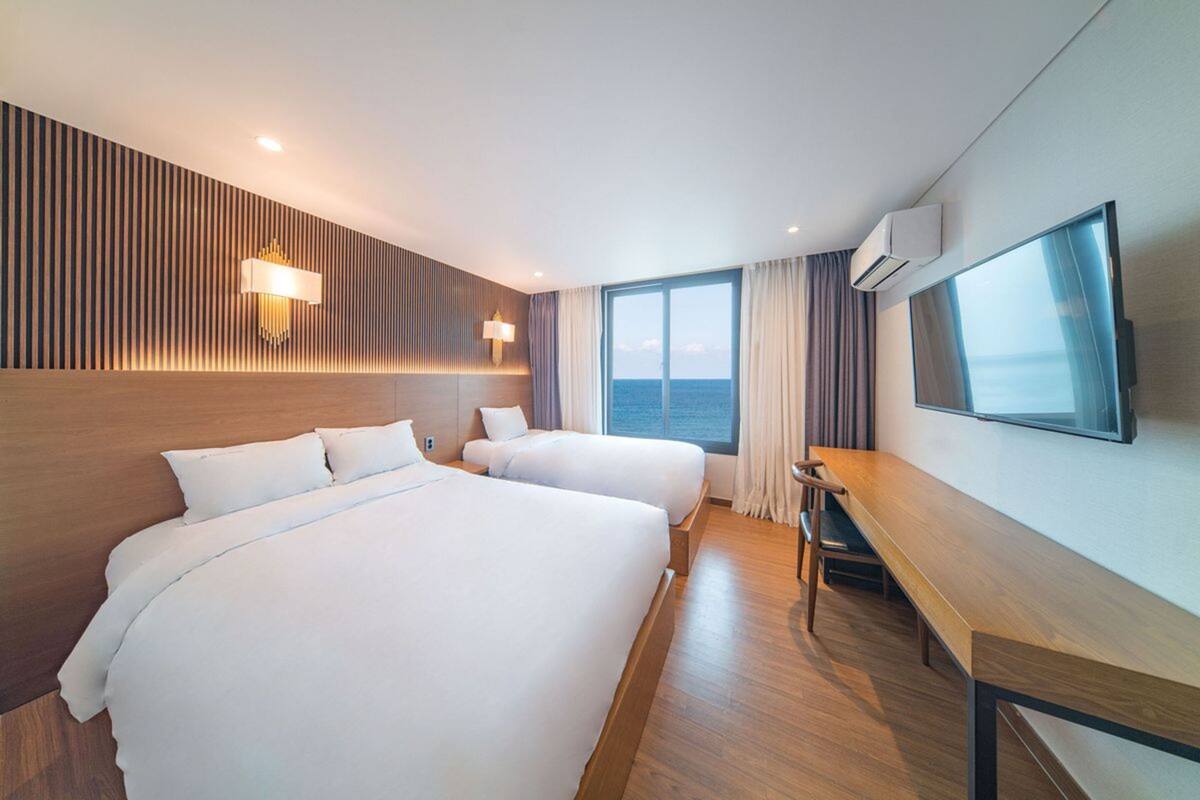 A hotel room with two beds, a desk, a TV, and a large window with a view of the sea.