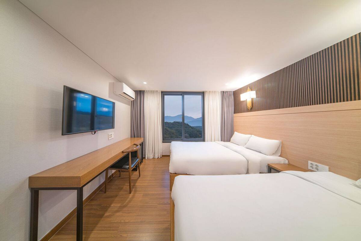 A hotel room with two beds, a desk, a TV, and a large window with a view of the mountains.