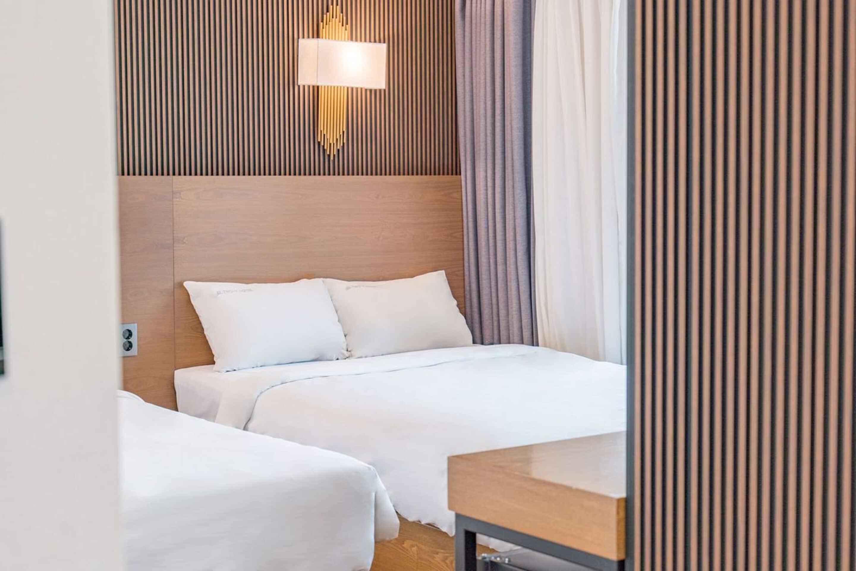 A hotel room with two beds, a wooden headboard, a bedside table, and a wall-mounted lamp.