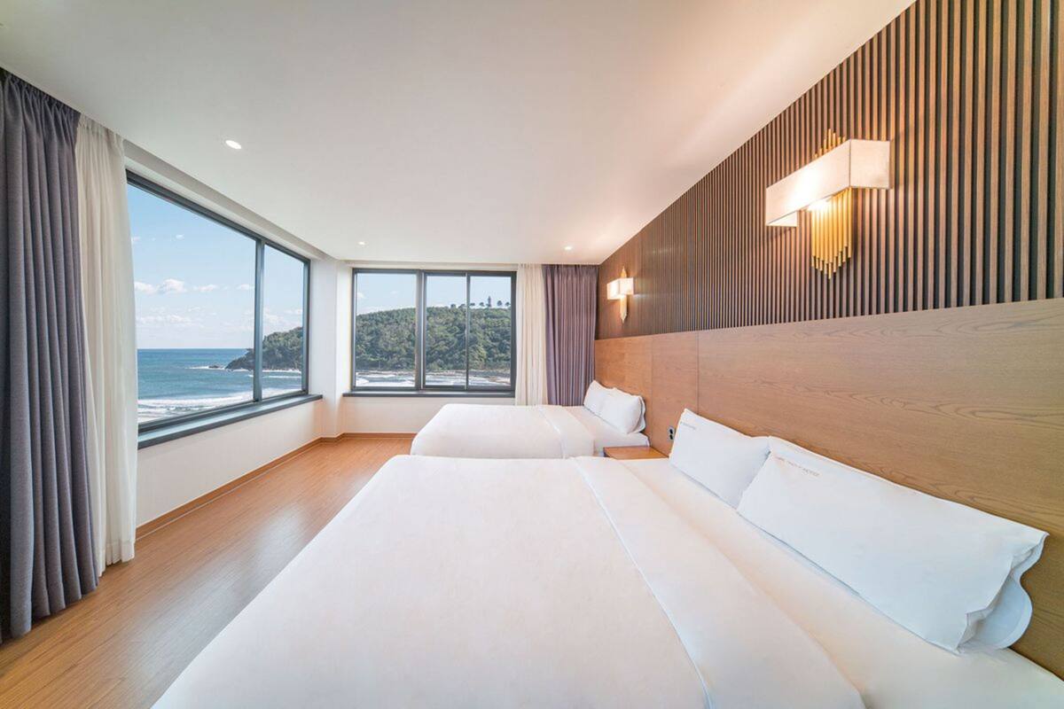 A hotel room with two beds, a large window with a view of the sea, and a wooden wall with a light fixture.