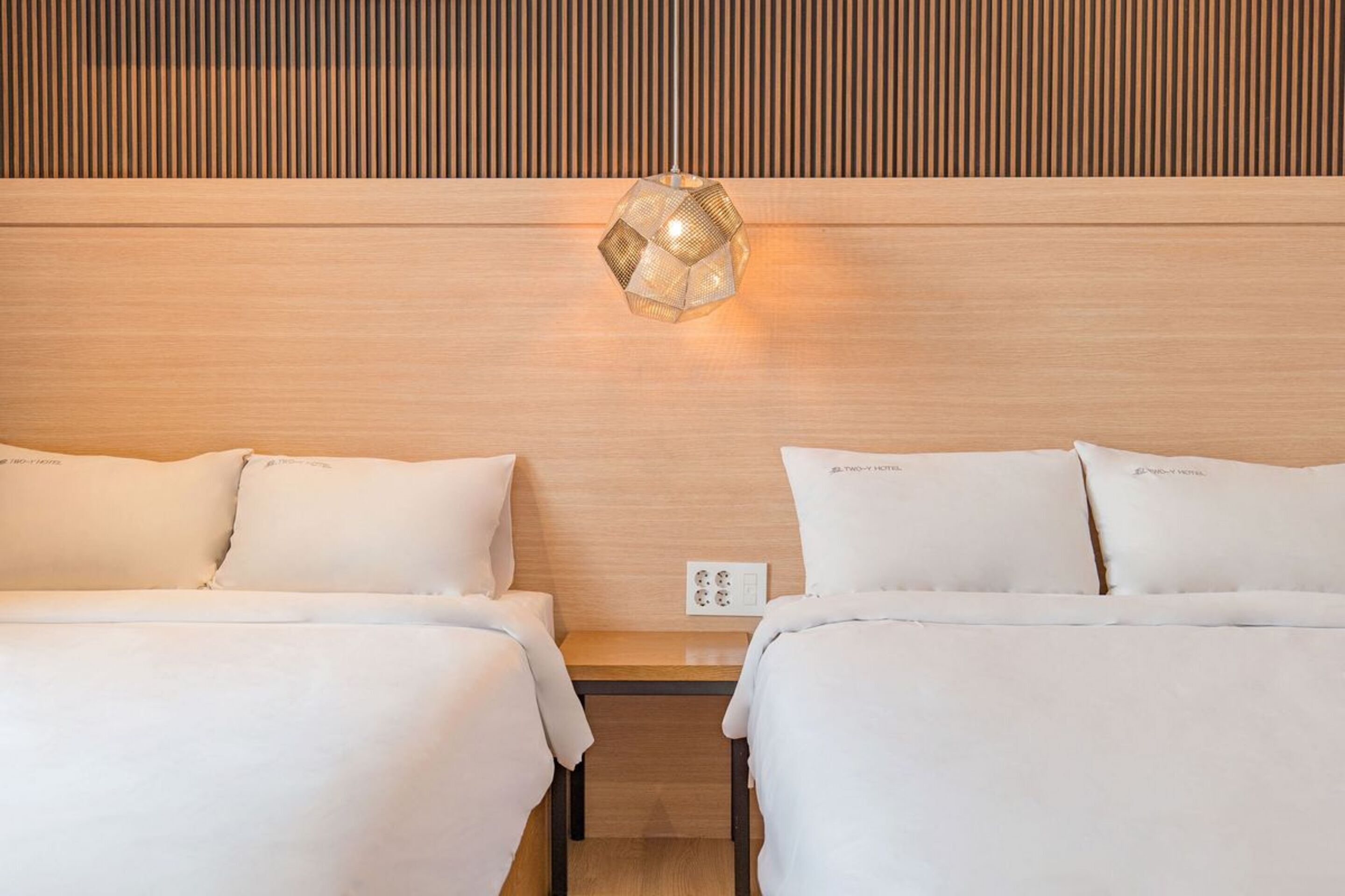 A hotel room with two beds, a wooden headboard, a wall-mounted light fixture, and a power outlet.