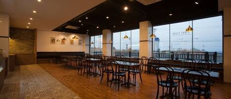 A spacious dining area with wooden flooring, multiple tables and chairs, and large windows offering a view of the cityscape.