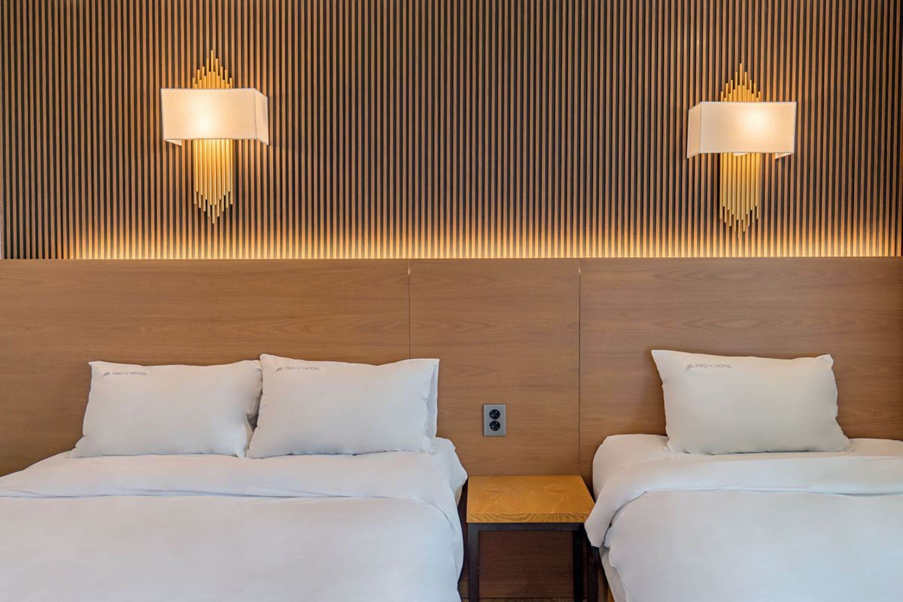 A hotel room with two beds, each with white bedding and pillows, and a wooden headboard with vertical slats. There are two wall-mounted lamps with a unique design.