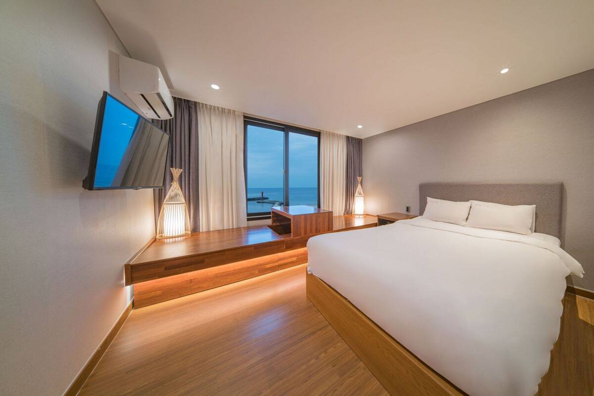 A modern hotel room with a large bed, a TV mounted on the wall, a desk with a chair, and a view of the sea through large windows.