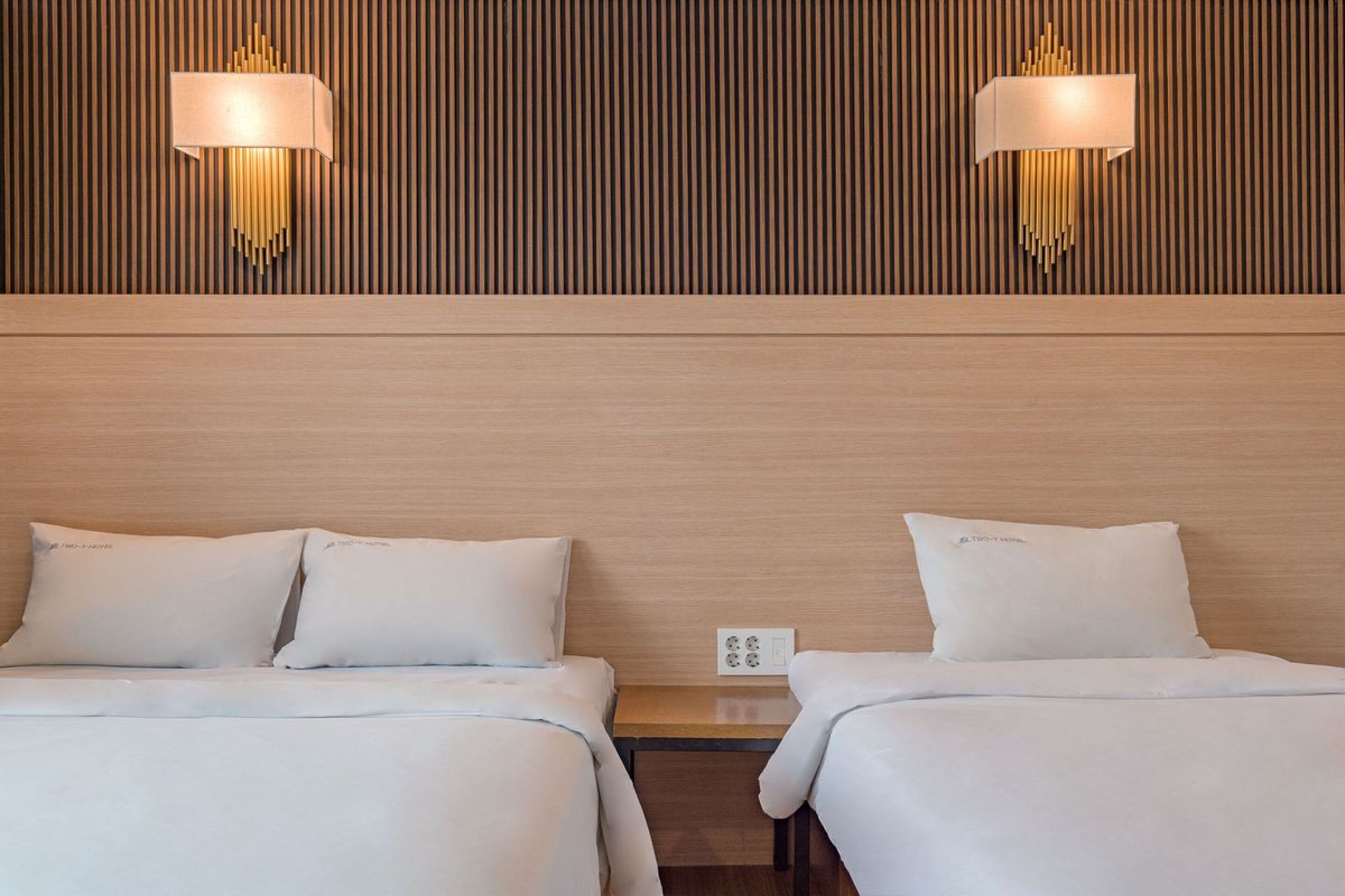 A hotel room with two beds, a wooden headboard, and wall-mounted lamps.