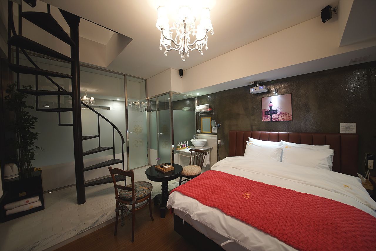 A hotel room with a bed, a chandelier, a glass-enclosed shower, a small table, and a chair.