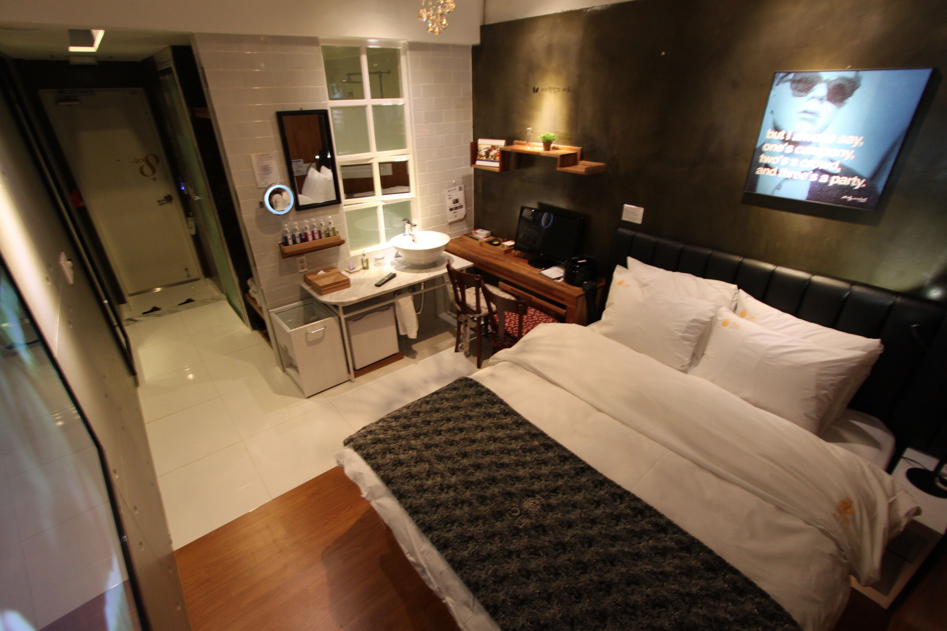 A modern hotel room with a large bed, a desk with a computer, a TV mounted on the wall, and a bathroom with a mirror and sink.