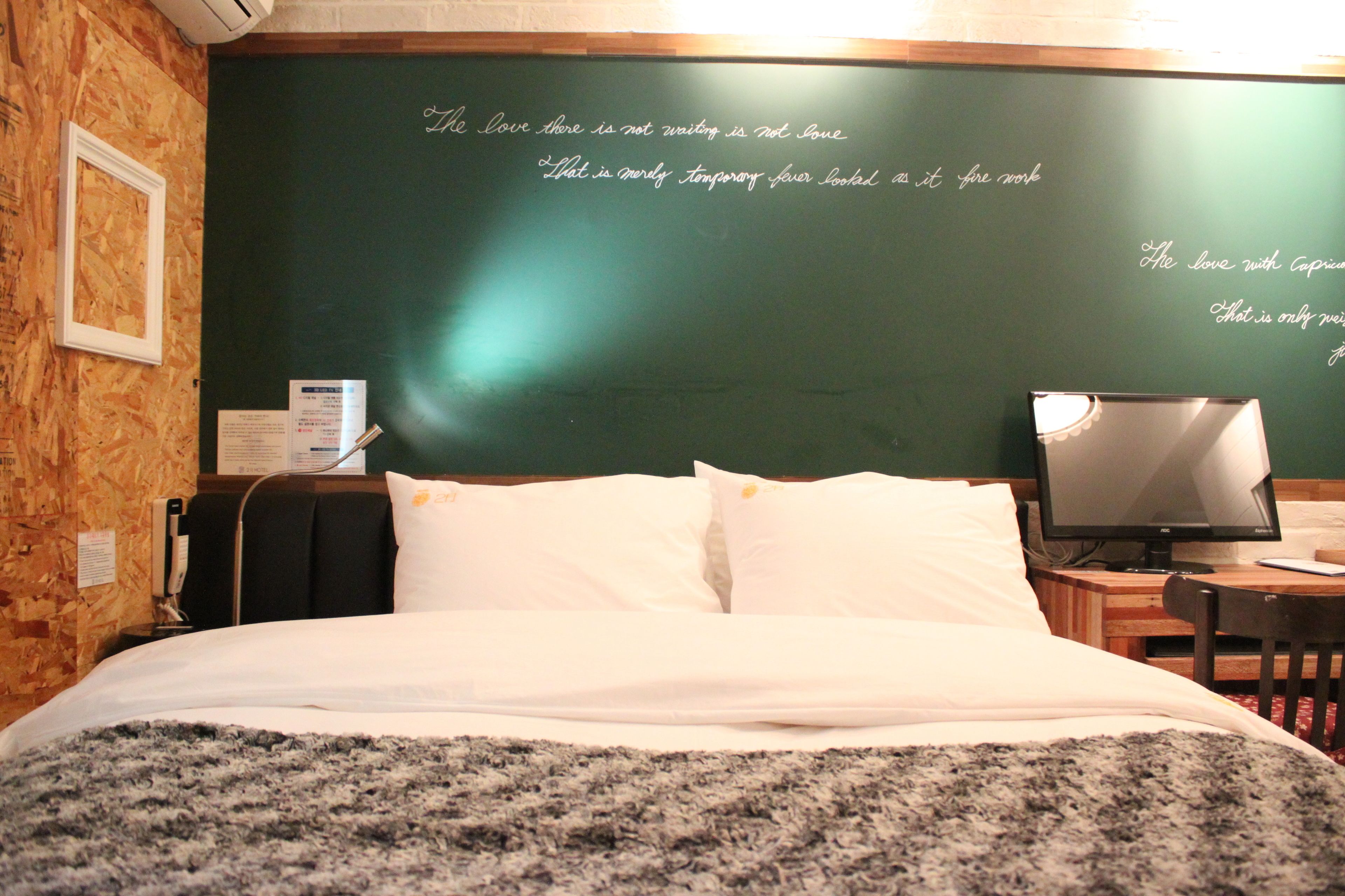 A neatly made bed with white linens and a gray blanket, a television on a wooden table, and a chalkboard wall with writing.