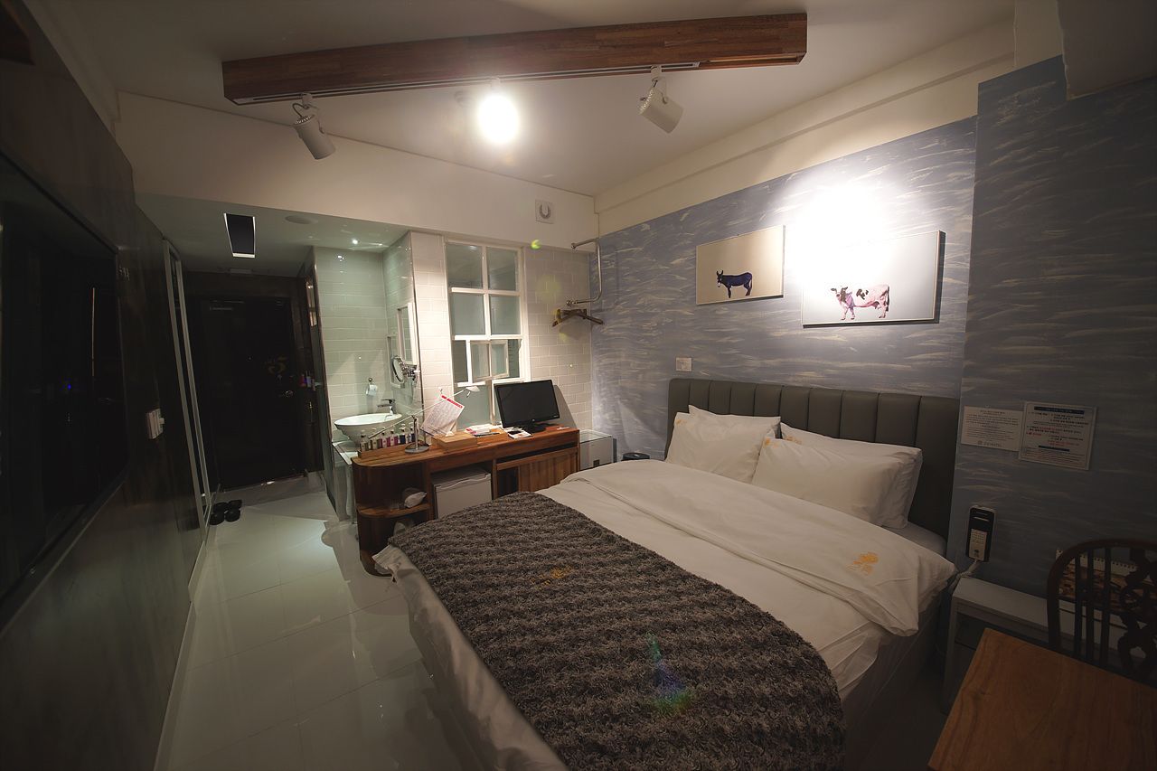 A modern hotel room with a bed, desk, and a bathroom visible through a glass door.