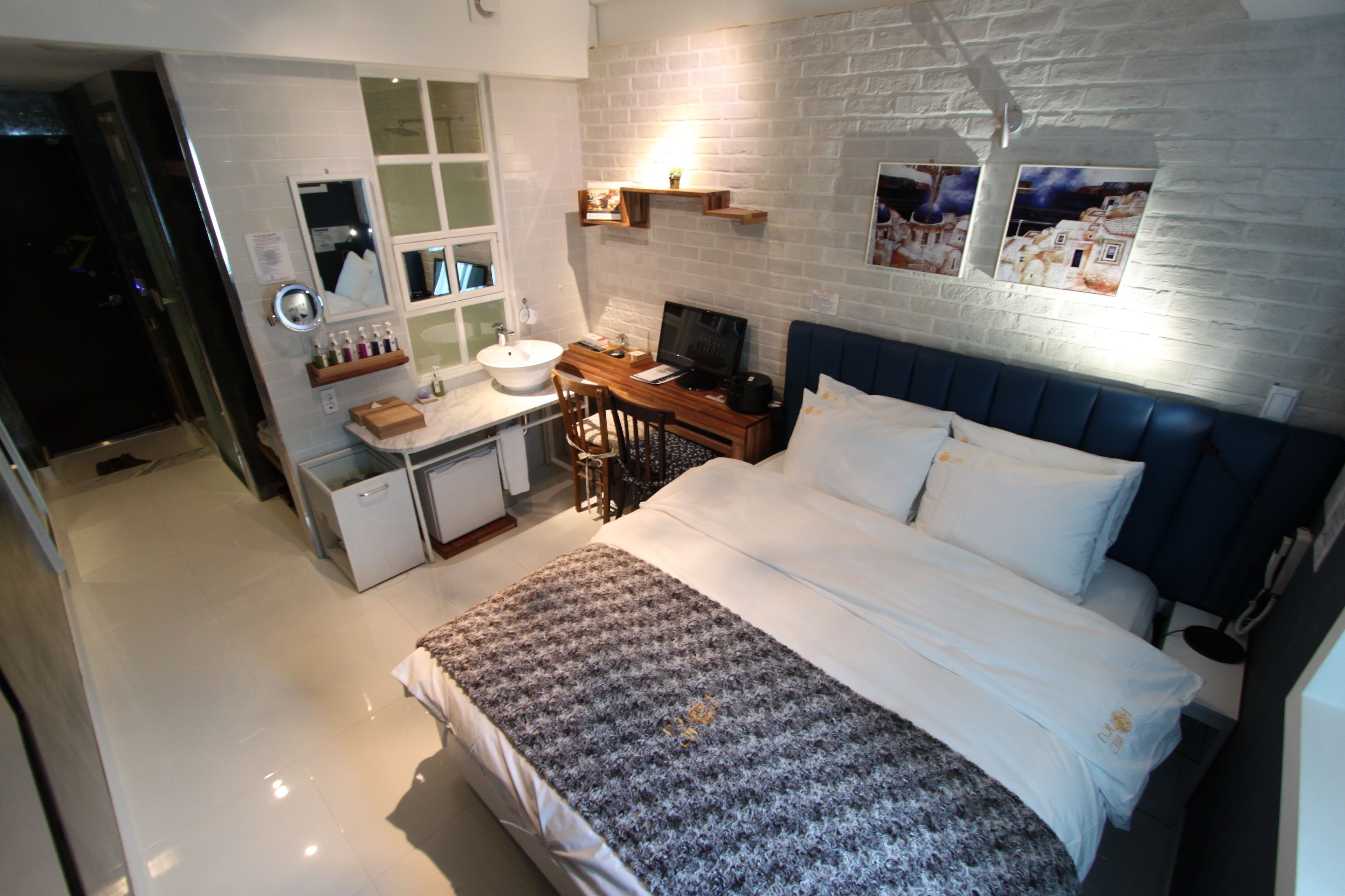 A modern hotel room with a large bed, a desk with a computer, a vanity with a mirror, and a bathroom with a sink and toilet.