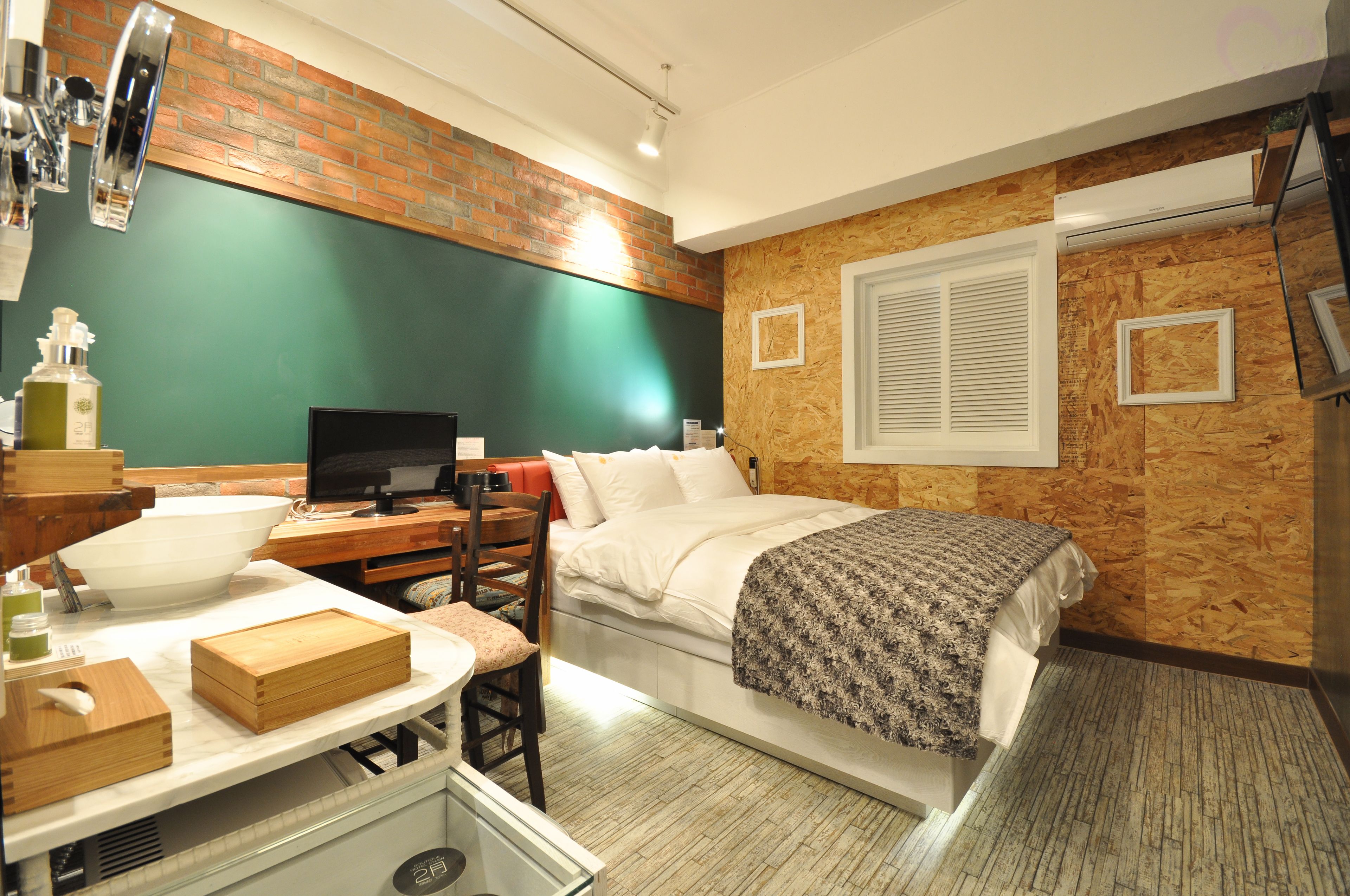A compact hotel room with a bed, a desk with a computer, a chair, and a small table with a bowl and a bottle.