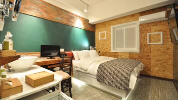 A compact hotel room with a bed, a desk with a computer, a chair, and a small table with a bowl and a bottle.
