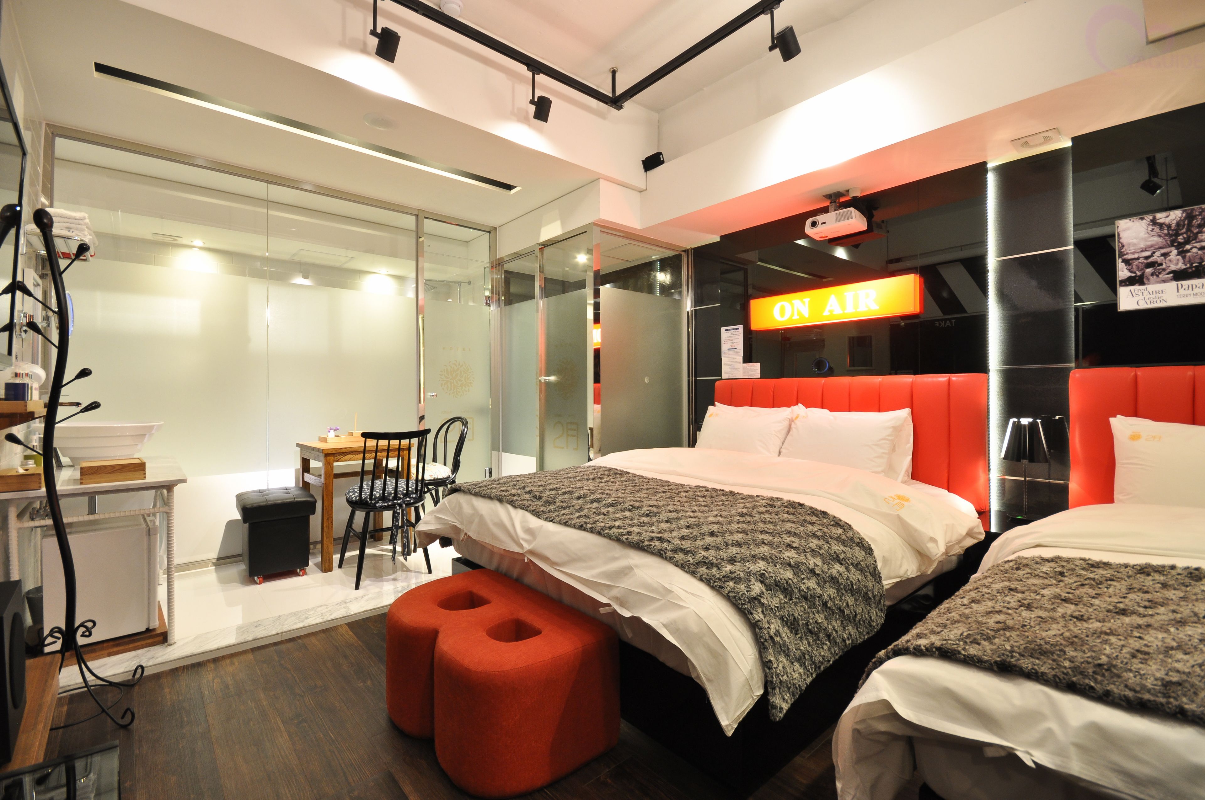 A modern hotel room with two beds, a red accent wall, a glass partition, a small table with chairs, and a sign that reads "ON AIR".