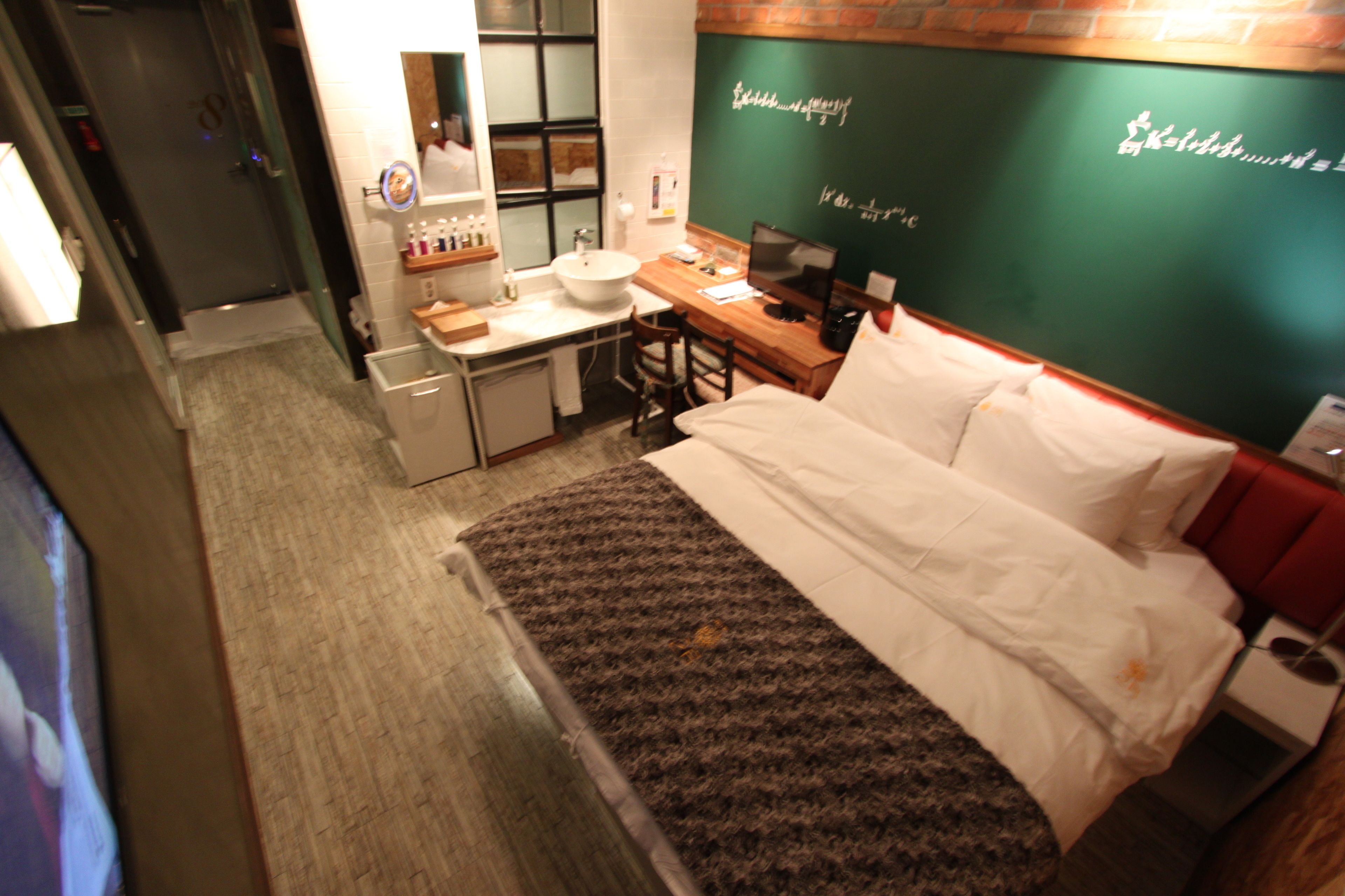 A hotel room with a bed, a desk with a computer, a sink, and a mirror.