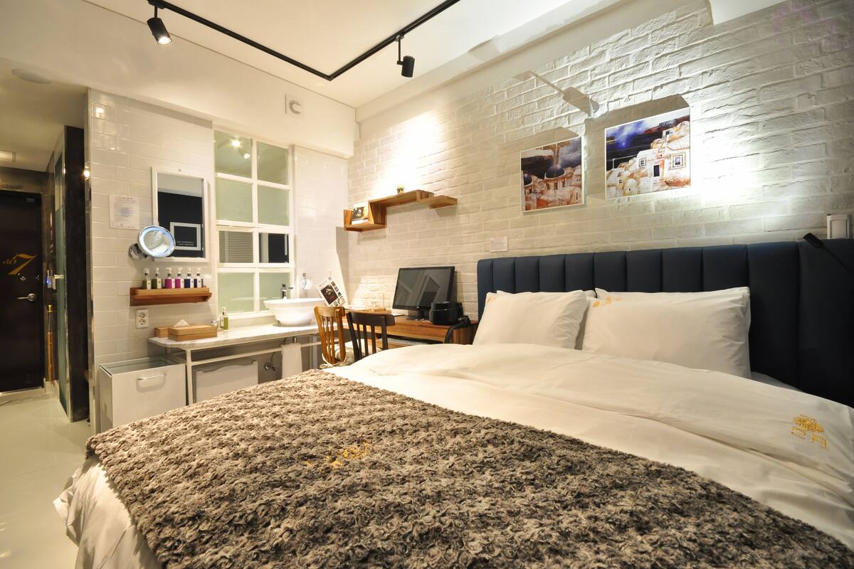 Ein modernes Hotelzimmer mit einem großen Bett, einem Schreibtisch mit Computer, einer kleinen Küchenzeile und einer Ziegelwand mit Kunstwerken.