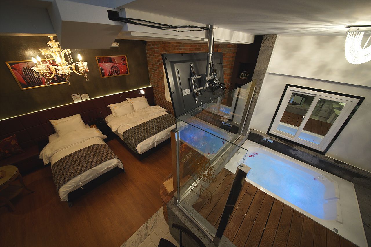 A modern hotel room with a glass-enclosed balcony, a large bed, a separate seating area, and a glass shower enclosure.
