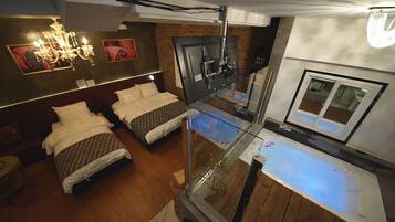 A modern hotel room with a glass-enclosed balcony, a large bed, a separate seating area, and a glass shower enclosure.