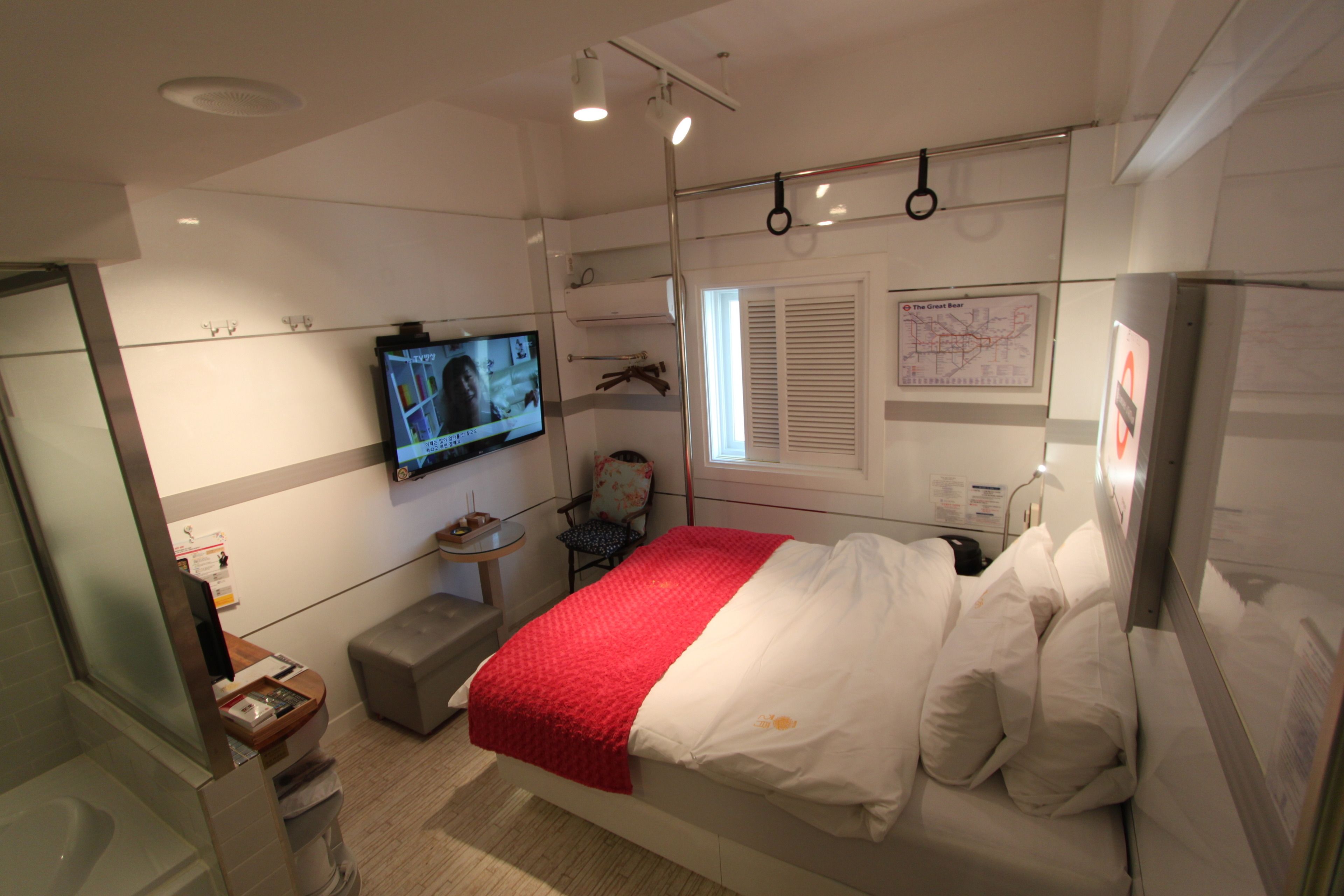 Habitació d'hotel moderna amb llit, televisió, taula petita i finestra amb persianes.