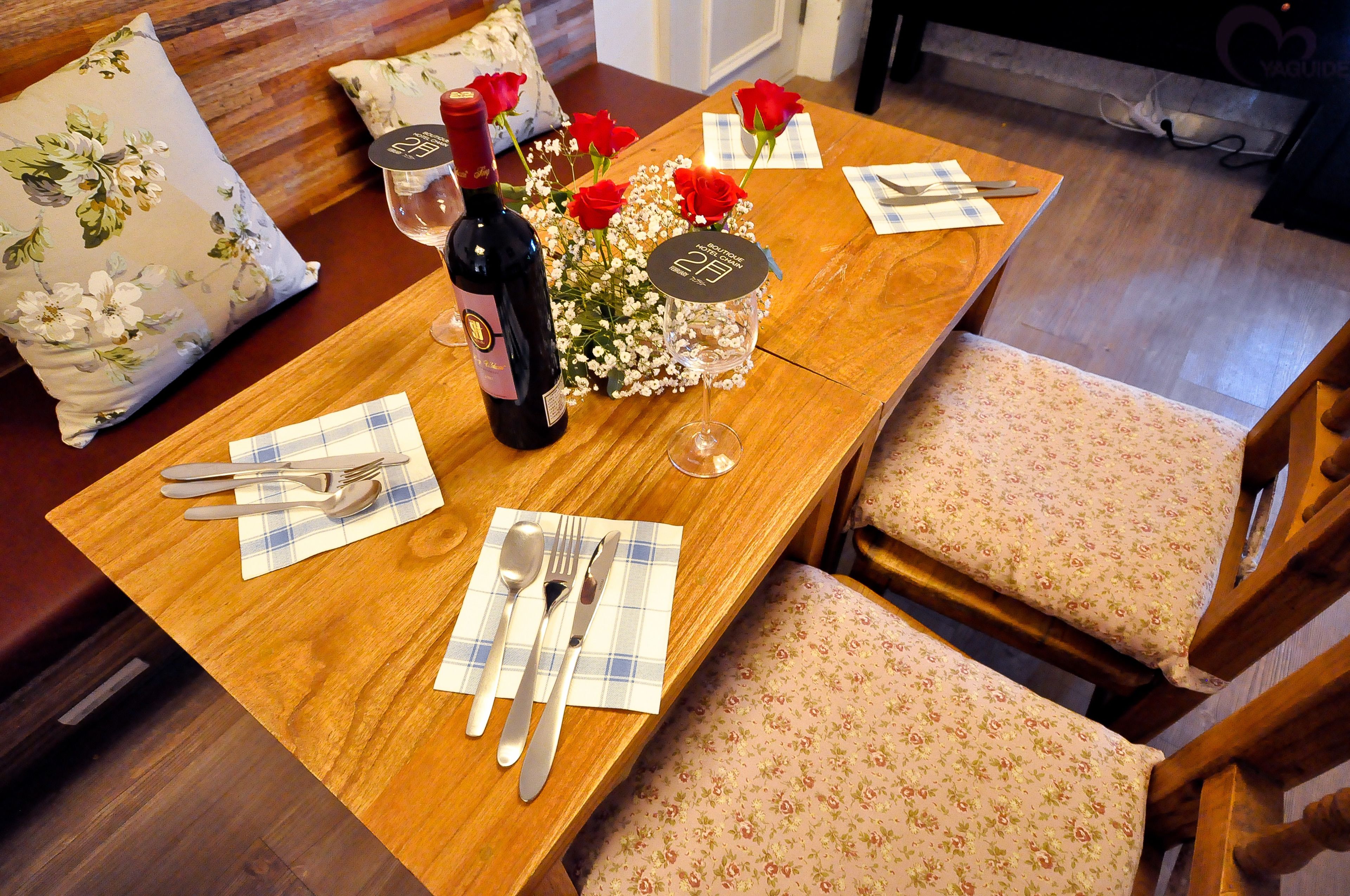 A wooden dining table set with floral cushions, a bottle of wine, and a bouquet of red roses.