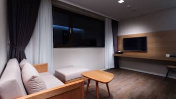 A modern hotel room with a wooden sofa, a small table, a TV mounted on the wall, and a large window with curtains.