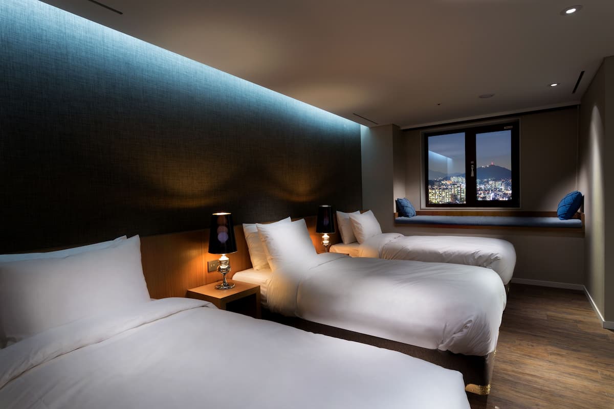 Habitación de hotel con dos camas, un ventanal grande con vistas a la ciudad y un banco junto a la ventana.