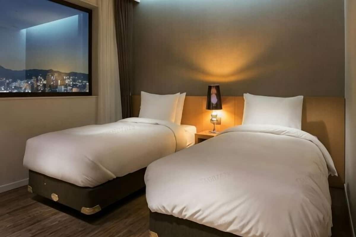 A hotel room with two beds, a nightstand, a lamp, and a window with a city view.