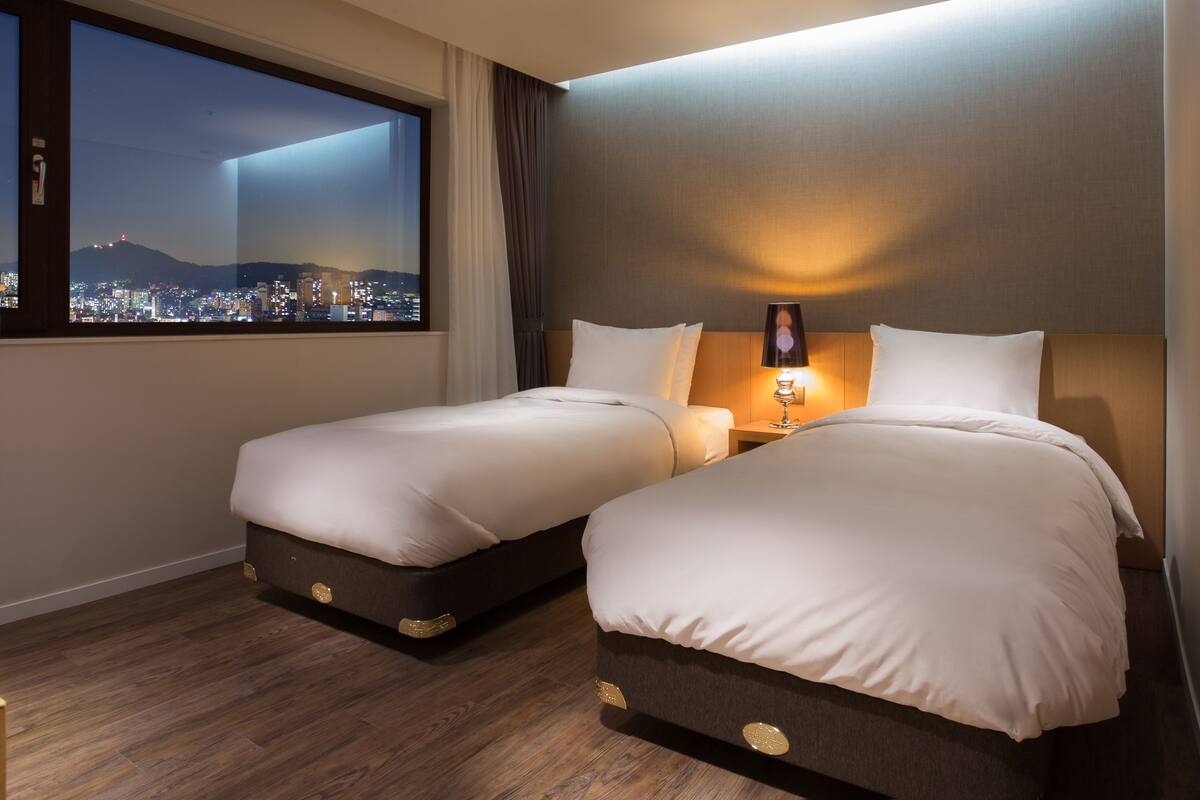Habitación de hotel con dos camas, una mesita de noche con lámpara, ventana con vista a la ciudad y suelo de madera.
