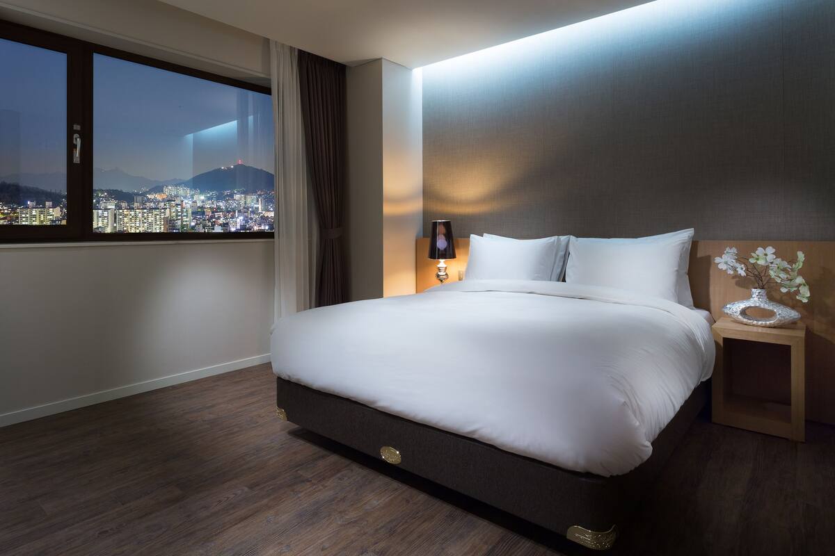 Una habitación de hotel moderna con una cama grande, una mesita de noche, una lámpara y vistas a un paisaje urbano a través de un amplio ventanal.